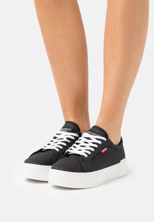Baskets noires en toile avec des lacets blancs et une épaisse semelle blanche. Dotées d'une étiquette de logo rouge sur le côté et d'un design de bout arrondi.