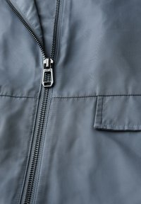 Veste grise avec un zip bien en vue, texture lisse et poche à l'avant. Le matériau semble léger et résistant à l'eau.