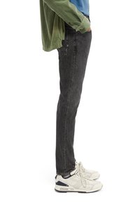 Jeans de mezclilla negra con un corte recto, combinados con una camisa de pana verde y zapatillas blancas con detalles en azul marino. Vista lateral.