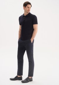 Granatowa koszulka polo z kołnierzykiem i trzema guzikami, zestawiona z ciemnymi spodniami i czarnymi butami z klamrą, stojąca na białym tle.