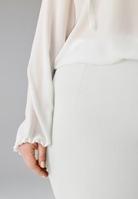 Blouse légère et transparente de couleur blanche avec des manches bouffantes, associée à une jupe ivoire lisse et ajustée. Texture subtile et détails de drapés doux.