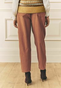 Roostevärvi ribikangas kitsenevad püksid tagataskutega, kantud koos kootud mustriga vestiga ja tumedate kontsakingadega puidust põranda taustal.