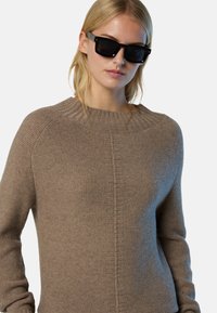 Brauner Strickpullover mit geripptem Ausschnitt, der ein strukturiertes Muster und eine lockere Passform aufweist. Er hat lange Ärmel. Schwarze Sonnenbrille getragen.