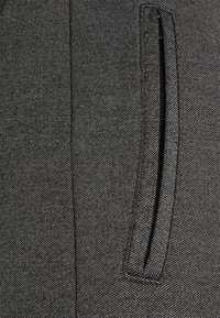 Zwarte textiele stof met een smalle, verticaal uitgelijnde zak met gestikte randen. Het oppervlak heeft een subtiel, fijn patroon.