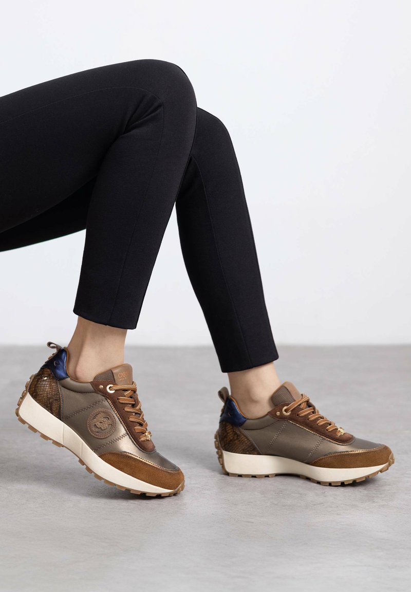 Braune und graue Sneaker mit strukturierten Akzenten und einer klobigen weißen Sohle. Sie verfügen über eine glatte Oberfläche, gemusterte Seitenpaneele und braune Schnürsenkel.