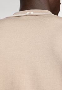 Smėlio spalvos, segmentuotas megztinis su minkšta tekstūra, pasižymintis kontrastinga balta apykakle ir subtiliais siuvimo akcentais.