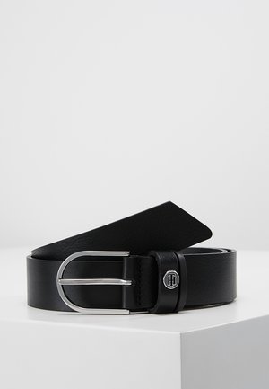 Ceinture en cuir noir avec boucle ovale argentée et petit emblème rond avec logo sur la boucle, présentée sur une surface blanche.