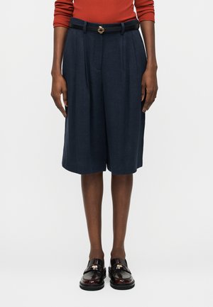 Άτομο που φοράει μπλε σκούρα πλισέ σορτς μέχρι το γόνατο με μαύρη ζώνη, μακρυμάνικη τοπ σε σκουριά απόχρωση και γυαλιστερά μαύρα μοκασίνια, στέκεται μπροστά σε απλό φόντο.