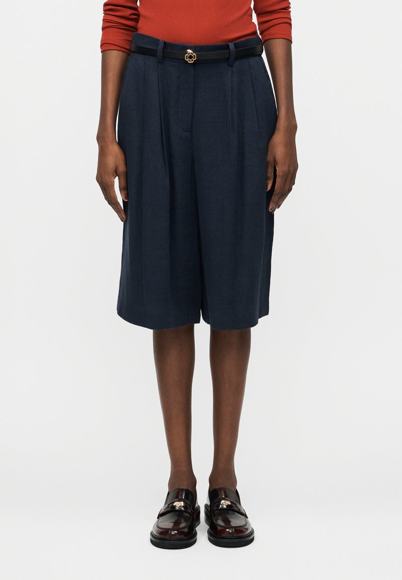 Άτομο που φοράει μπλε σκούρα πλισέ σορτς μέχρι το γόνατο με μαύρη ζώνη, μακρυμάνικη τοπ σε σκουριά απόχρωση και γυαλιστερά μαύρα μοκασίνια, στέκεται μπροστά σε απλό φόντο.