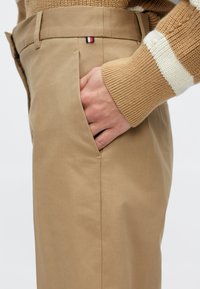 Bež pamučne hlače s bočnim džepom, s malom crveno-bijelo-plavom oznakom logotipa na pojasu. Tkanina ima glatku teksturu.