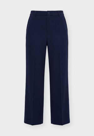 Femme portant un pull zippé en maille torsadée bleu marine, un pantalon large assorti et des baskets blanches, debout devant un fond clair.