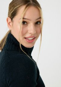 Suéter de cuello alto de punto texturizado en verde oscuro con detalles acanalados en el cuello, llevado por un modelo con cabello lacio y claro.