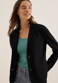 Femme portant un blazer noir sur un débardeur teal et un jean gris, regardant légèrement sur le côté avec les cheveux lâches et un fond neutre.