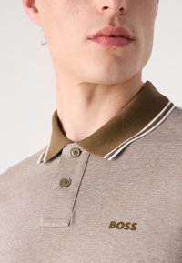 Ruda polo marškinėliai su kontrastiniu ruduo apykaklės, dviem mygtukais ir baltais dryžiais apykaklės krašte; turi logotipą "BOSS" rudai.