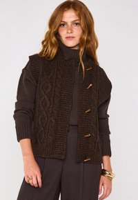 Gilet lavorato a maglia marrone con un pannello frontale testurizzato, dotato di bottoni in legno, colletto alto e maniche a costine. Indossato sopra un dolcevita coordinato.