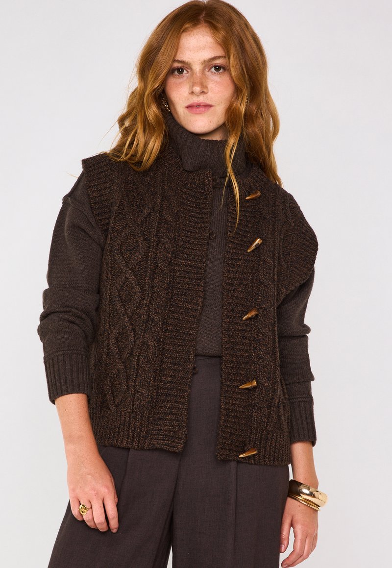 Gilet lavorato a maglia marrone con un pannello frontale testurizzato, dotato di bottoni in legno, colletto alto e maniche a costine. Indossato sopra un dolcevita coordinato.
