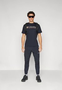 T-shirt preta com o logótipo da Columbia, calças leves pretas e uma mochila preta. Os acessórios incluem óculos de sol escuros e calçado preto.