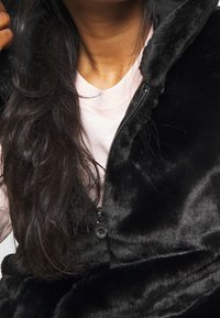 Black faux fur vest with a zip-up front, featuring a smooth texture and a detachable hood. Worn over a light pink shirt.
