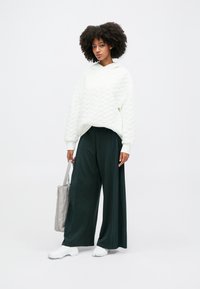 Felpa bianca, con texture e cappuccio, decorata con un motivo ondulato, abbinata a pantaloni larghi plissettati di un verde scuro e a calzature bianche. Porta con sé una borsa tote argentata.