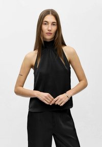 Schwarzes Satin-Haltertop mit hohem Kragen, lockerer Passform und glatter Textur. Getragen mit passenden schwarzen Hosen posiert das Modell mit verschränkten Händen.
