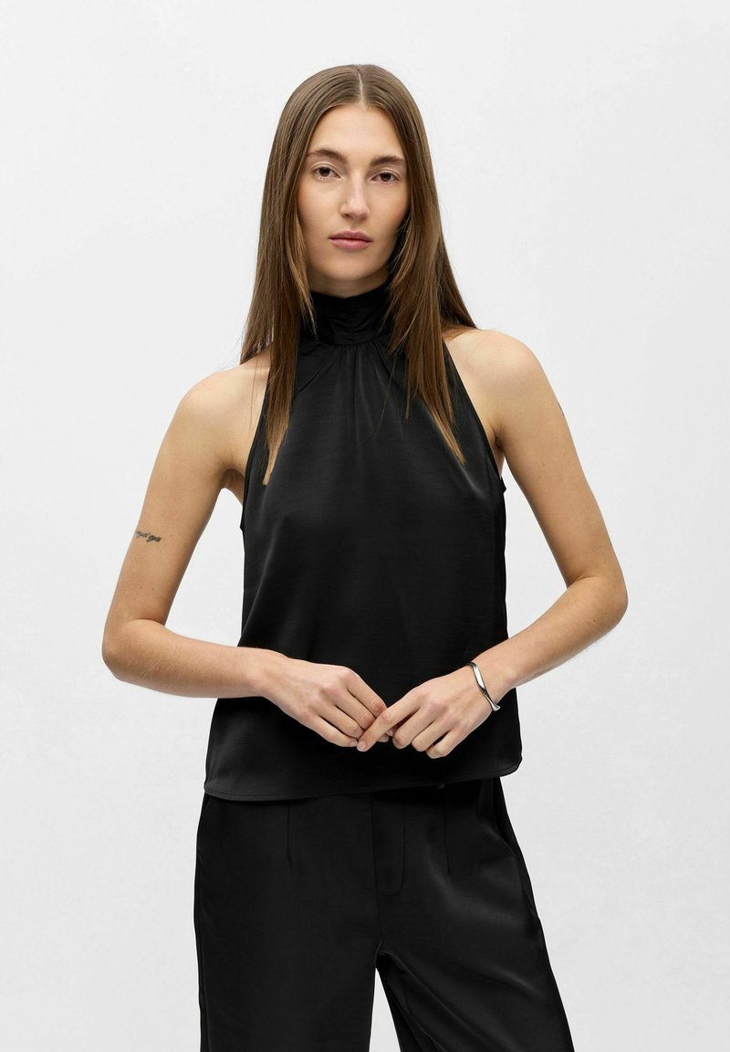 Sort satin halterneck top med høj hals, afslappet pasform og glat tekstur. Båret med matchende sorte bukser, poserer modellen med armene korslagt.