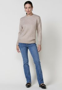 Suéter de punto beige con un patrón texturizado, cuello redondo y puños acanalados, acompañado de jeans de mezclilla azul claro y zapatos planos negros.