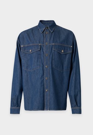 Jeune homme portant une veste en jean sombre et ample, un pantalon large à rayures verticales, et des chaussures noires avec des bouts dorés, debout devant un fond uni.