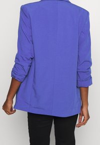 Osoba v purpurovém sakě s ohrnutými rukávy stojí proti jednobarevnému pozadí, zobrazuje zadní část oblečení.