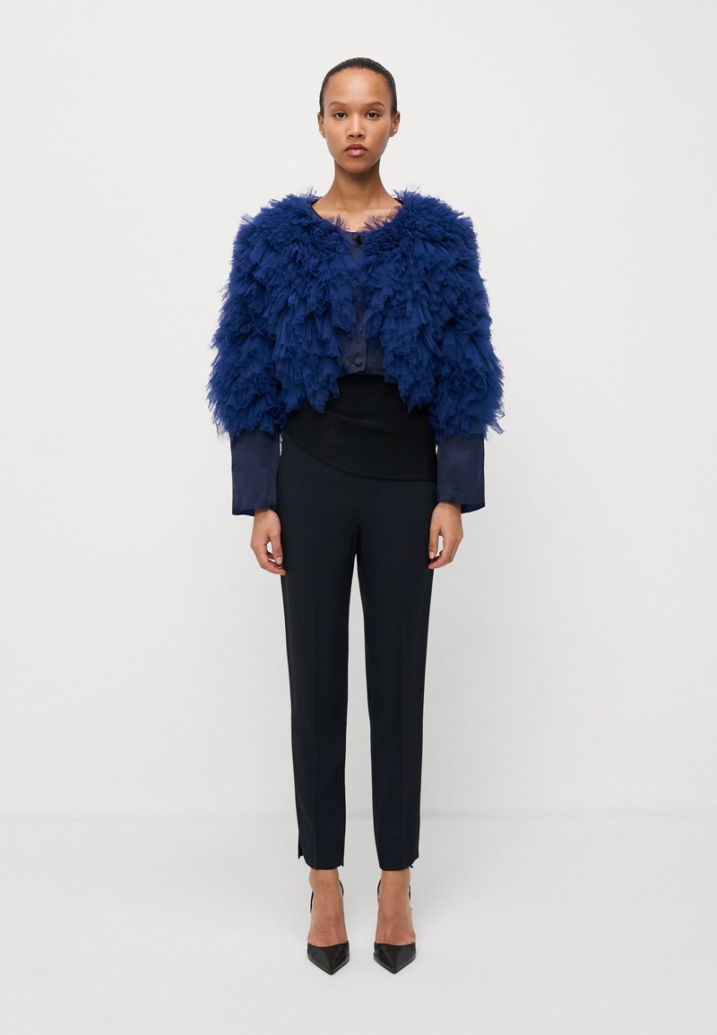 Navy blue' kurtka o krótkim kroju z teksturą piór z okrągłym dekoltem, zestawiona z eleganckimi czarnymi spodniami i szpiczastymi czarnymi szpilkami.