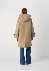 Beige wasserabweisender Mantel mit Kapuze, breiten Ärmeln und einem einzelnen Knopfdetail an den Bündchen, kombiniert mit blauen Schlagjeans.