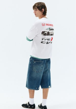 Jeune homme debout, les bras croisés, portant un t-shirt blanc avec un graphisme Honda Civic Type R, un short en jean long, des chaussettes blanches et des baskets noires.