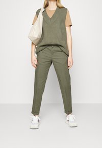 Olivově zelená pletená vesta přes béžové tričko, doplněná olivově zelenými kalhotami a bílými teniskami. Celkový vzhled dokončuje krémová kabelka.