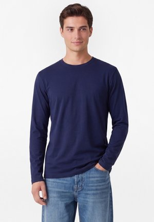 Giovane uomo con una maglietta blu navy semplice a maniche lunghe e jeans azzurri, in piedi con una mano in tasca, guarda leggermente di lato.