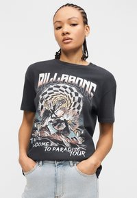 Γυναίκα με πλεγμένα μαλλιά που φοράει σκούρο T-shirt της Billabong με σχέδιο λουλουδιών, καρό σπείρα, βουνά και κείμενο "Καλώς ήρθατε στην περιοδεία Paradise."