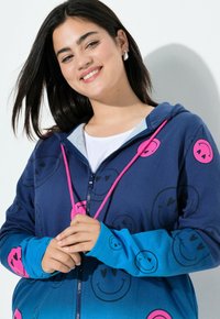 Sudadera con cremallera de color azul marino con patrones de caritas sonrientes en rosa, puños azules en contraste y cordones rosa. Tejido suave con efecto degradado.