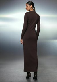 Frau mit dunklem Pferdeschwanz, die ein enganliegendes, langärmliges braunes Maxikleid und braune gemusterte High-Heels-Stiefel vor einem grau verlaufenden Hintergrund trägt.