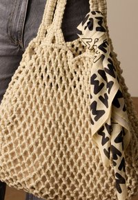 Beige haak-tote bag met dikke hengsels, voorzien van een gestructureerd gaaspatroon en versierd met een lichte, gestreepte sjaal in zwart en crèmekleur.