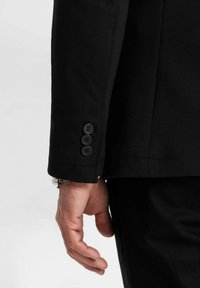 Närbild av en persons vänstra hand bredvid ärmen på en svart kavaj med tre knappar, bärande en silverfärgad armbandsur.