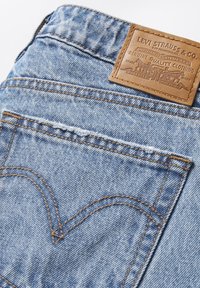 Jean en denim bleu avec une étiquette en cuir embossée du logo. La couture notable comprend un motif courbé sur la poche arrière.