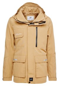 Giubbotto impermeabile di colore beige con cappuccio, diverse tasche, chiusura con cerniera e dettagli riflettenti. Tessuto liscio con cuciture decorative.
