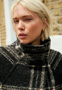 Manteau en laine à carreaux noir et blanc avec un col haut et un tissu texturé, présentant un motif de carreaux marqué et une finition douce.