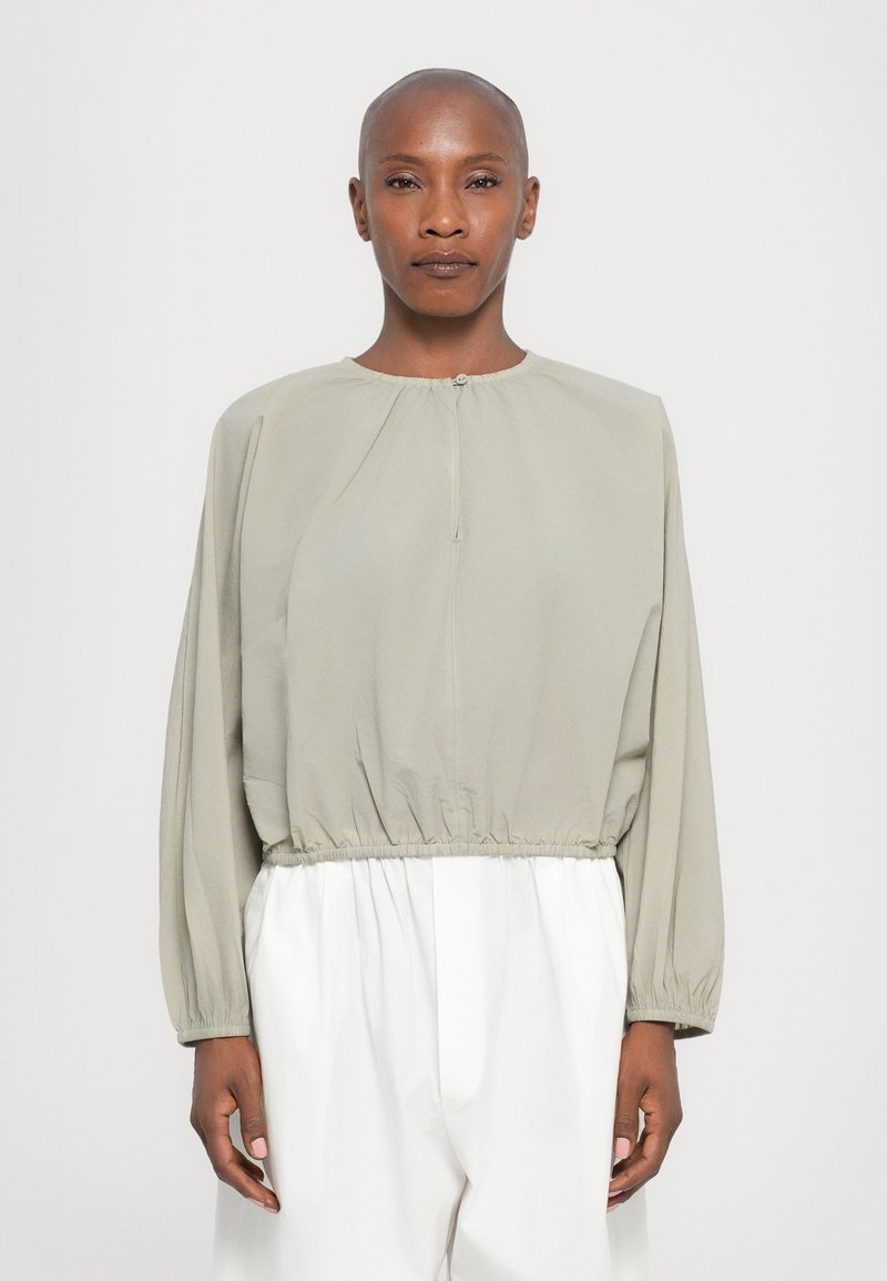 Blusa verde oliva chiaro con maniche lunghe, vita elastica e collo alto; texture liscia con dettagli di raccolta sottili.