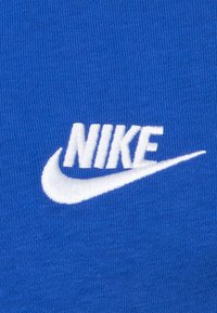 Modré textilní pozadí s bílým vyšívaným logem „Nike“ a motivem šipky, s hladkou texturou a jasným kontrastem.