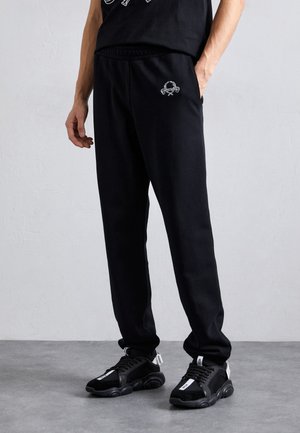 Black sweatpants with an elastic waistband, featuring a small embroidered logo. Paired with black sports shoes, highlighting a textured design.