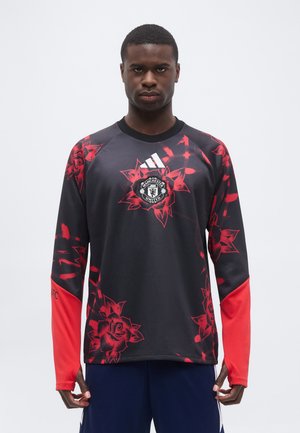 Muž nosí čierno-červený futbalový dres Manchester United s kvetinovým vzorom a logom Adidas na hrudi.