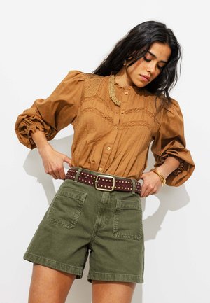 Femme ajustant une ceinture marron cloutée portée sur un short vert taille haute et un chemisier marron à manches longues avec des détails texturés.