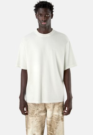 Giovane uomo con pelle scura e capelli corti ricci, indossa una maglietta bianca larga e pantaloni marroni chiaro con trama, in piedi fermo.