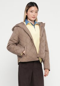 Klädd med en quiltad brun jacka med huva, med en framsida som har dragkedja och sidofickor, lager på lager över en gul stickad topp och en blå skjorta med krage.