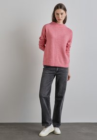 Roze gebreide trui met een ribbeltextuur, voorzien van een hoge halslijn en lange mouwen, gecombineerd met donkergrijze rechte jeans en witte sneakers.