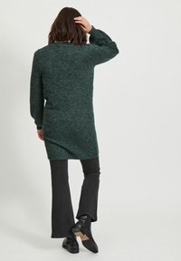 Maglione verde lavorato a maglia con maniche lunghe e orlo asimmetrico, abbinato a jeans neri a zampa e stivali neri. Aspetto morbido e texturizzato.